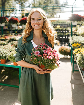 Floristin Paula steht mit einem strahlenden Lächeln in ihrem Gartencenter und hält einen Blumentopf mit Blumen in der Hand